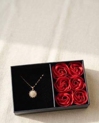 Caja de regalo abierta con el collar girasol dorado y seis rosas rojas sobre lino crema