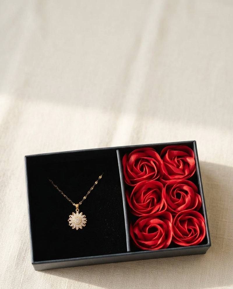 Caja de regalo abierta con el collar girasol dorado y seis rosas rojas sobre lino crema